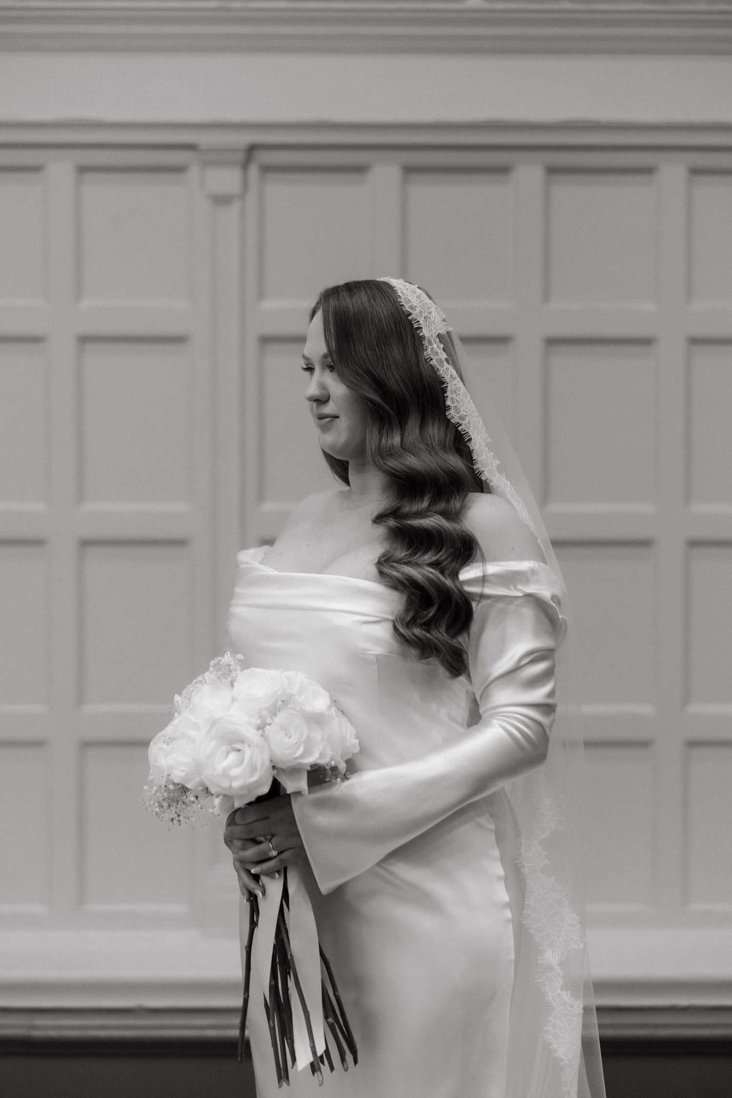 Black and white photo of a bride in an off-shoulder gown holding a white rose bouquet, wearing a lace veil.