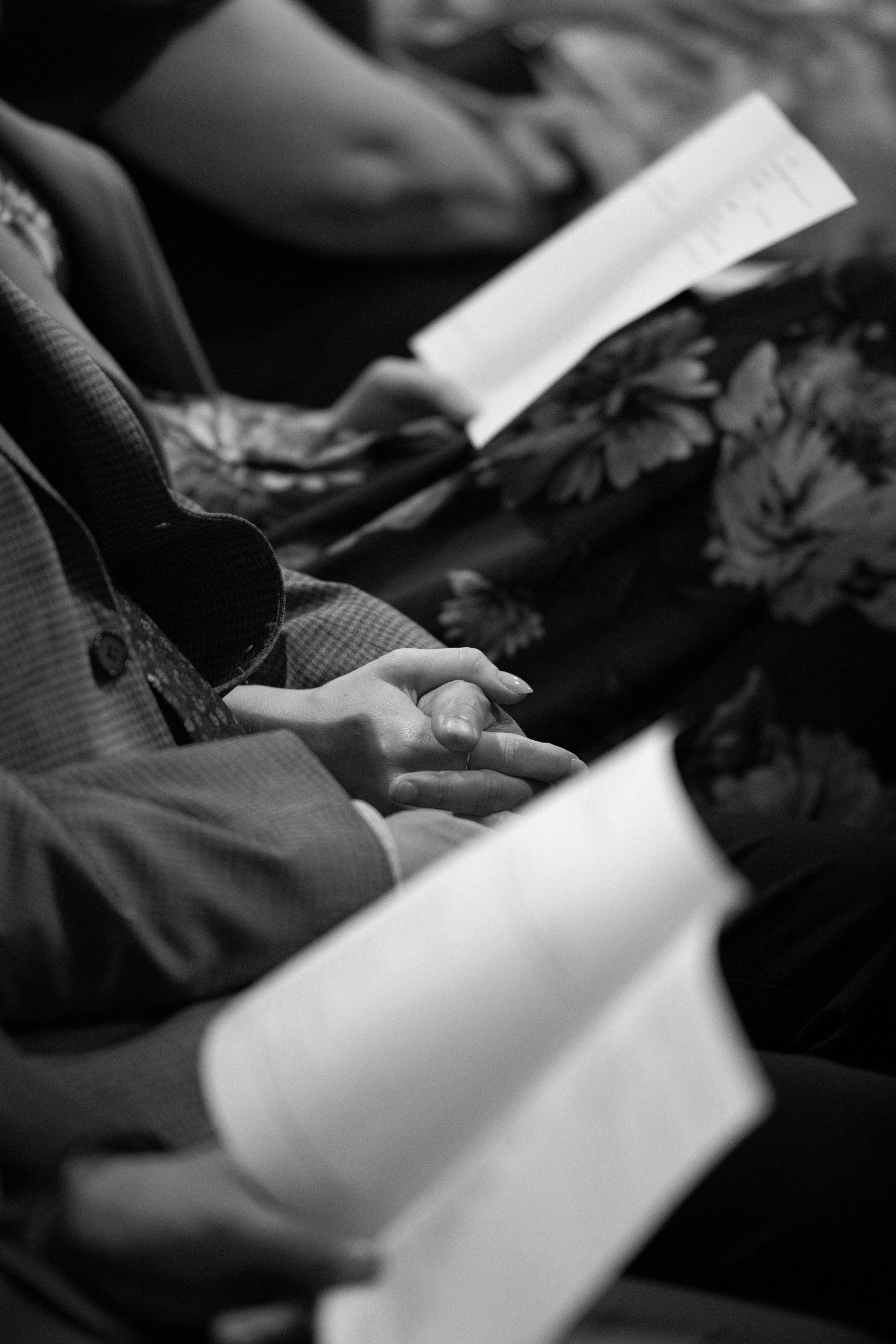 Black and white photo of people sitting holding open books or programs, with clasped hands visible in the foreground.