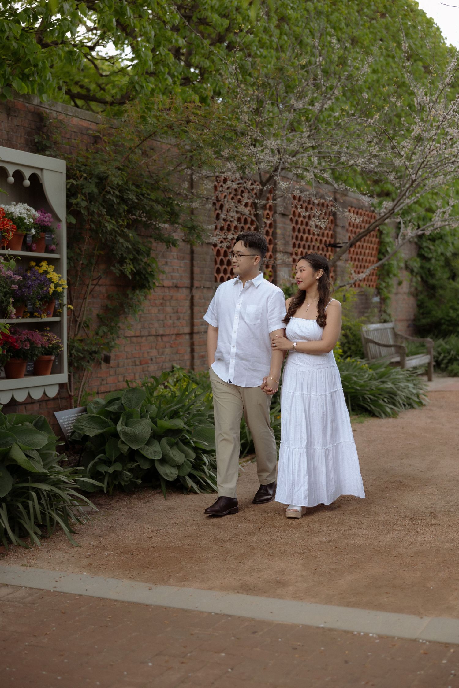 Couple in white outfits holding hands in a garden pathway with brick walls and colorful flowers.