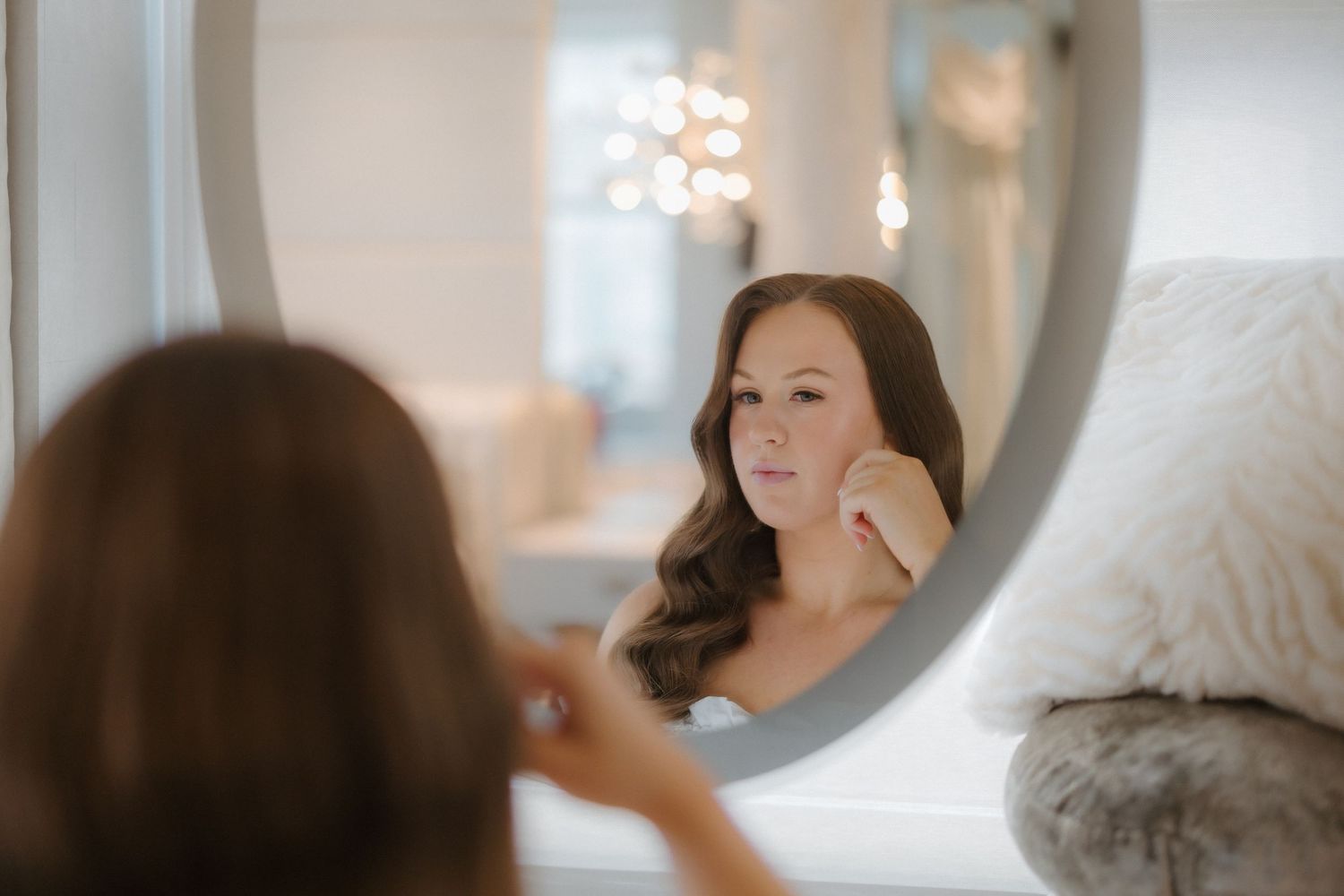 Woman with wavy hair gazes at her reflection in a round mirror while touching her face in an elegant room.