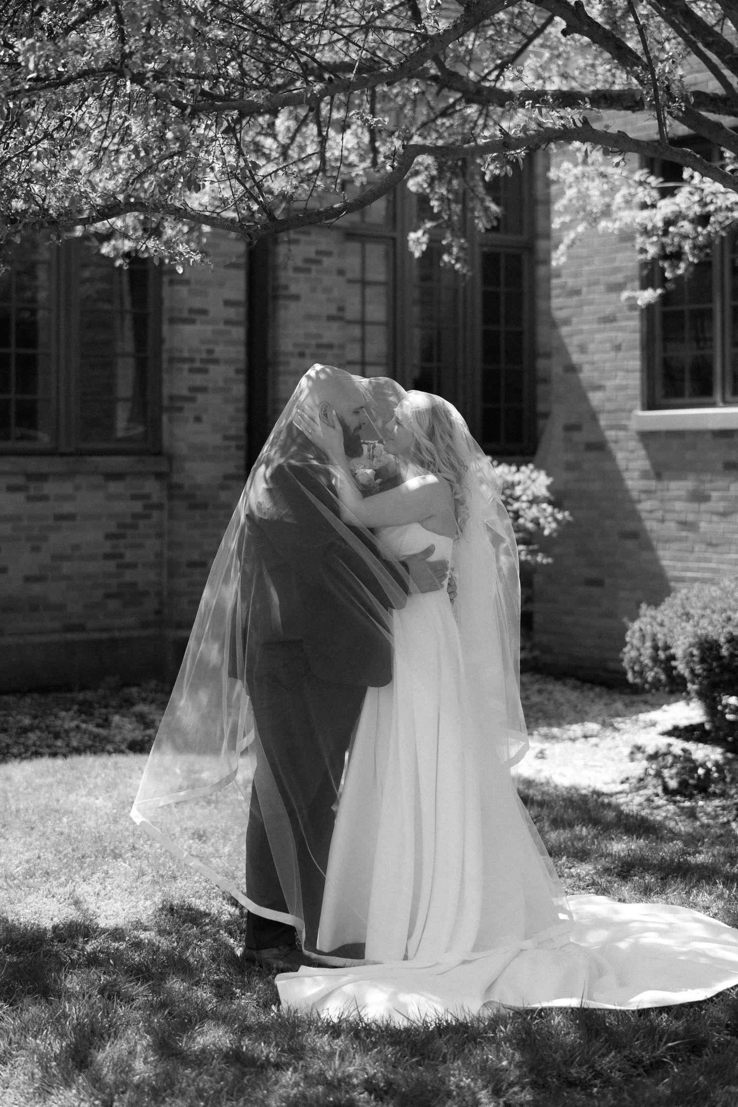 Black and white photo of bride and groom kissing under veil outside brick church on wedding day.