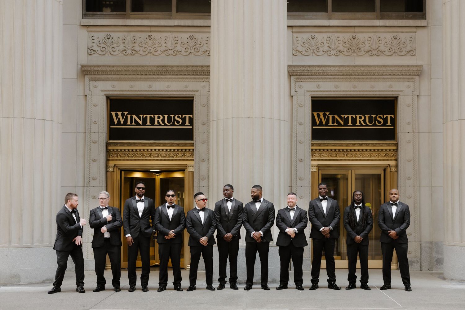 Groomsmen in black tuxedos posing in front of Wintrust building with gold signage in Chicago.