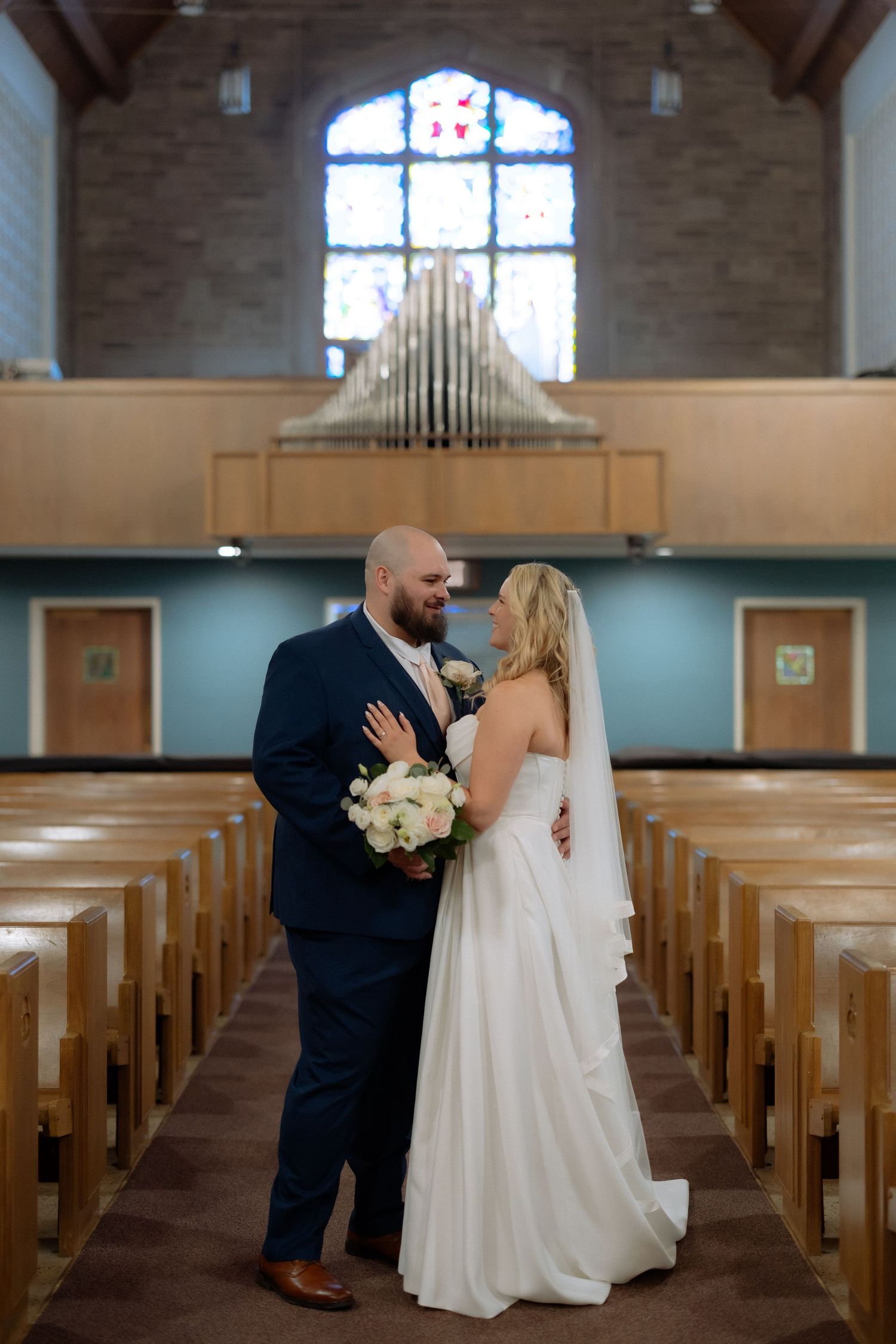 Bride and groom share a romantic moment in a church aisle, with stained glass windows and wooden pews in the background.