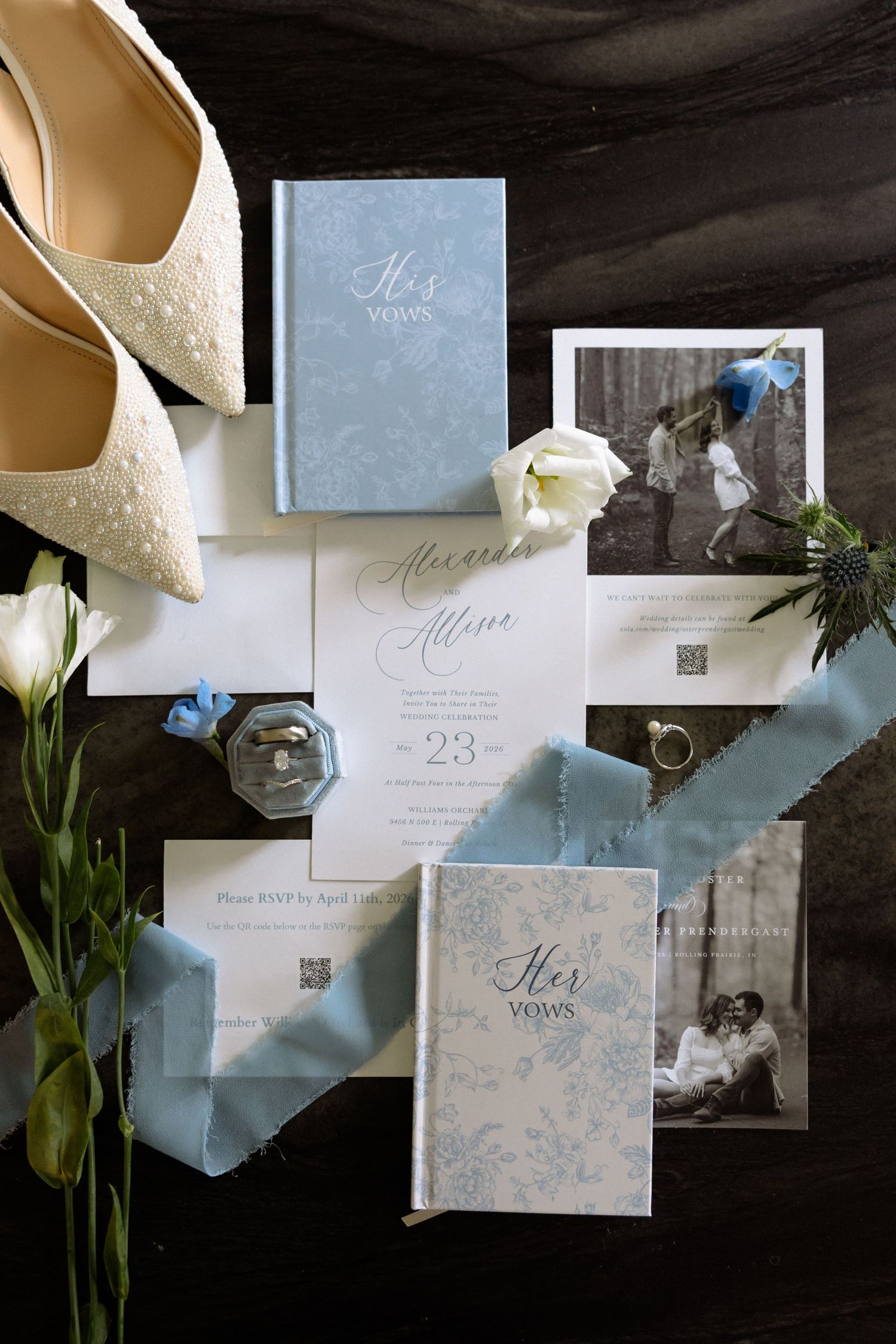 Wedding flat lay with dusty blue invitations, vow books, rings, and bridal heels on dark surface.