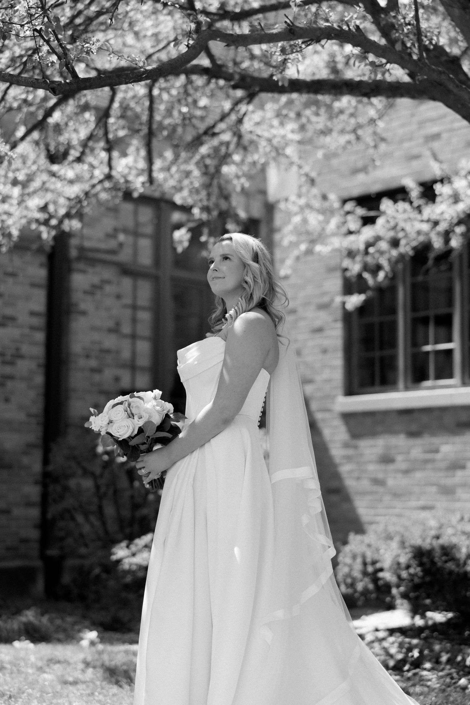 Elegant bride in a flowing white gown holding a bouquet, standing under blossoming trees.