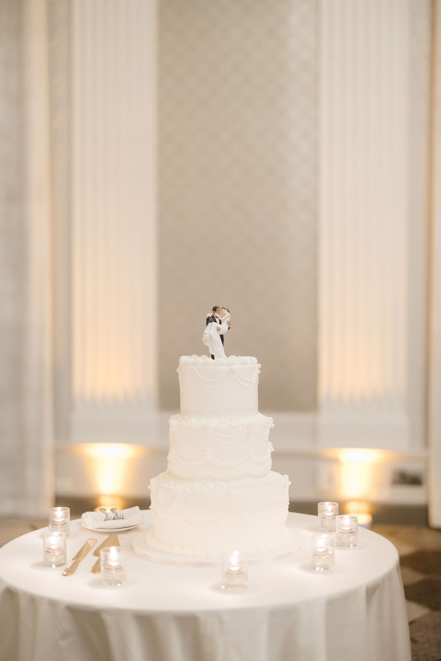 Elegant three-tier white wedding cake with bride and groom topper, surrounded by candles on a draped table.