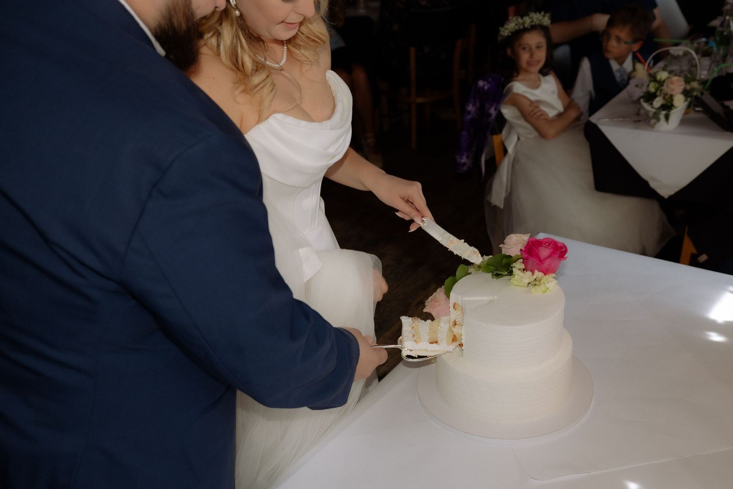 Bride and groom cutting a white two-tier wedding cake decorated with a pink rose at their wedding reception.