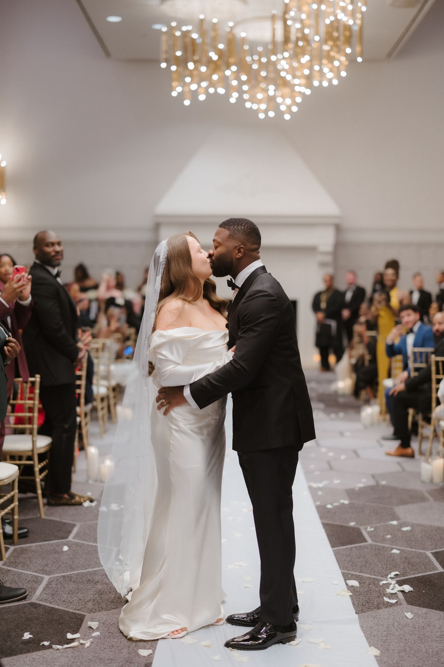 Interracial couple shares first kiss at elegant wedding ceremony with gold chandeliers and seated guests applauding.