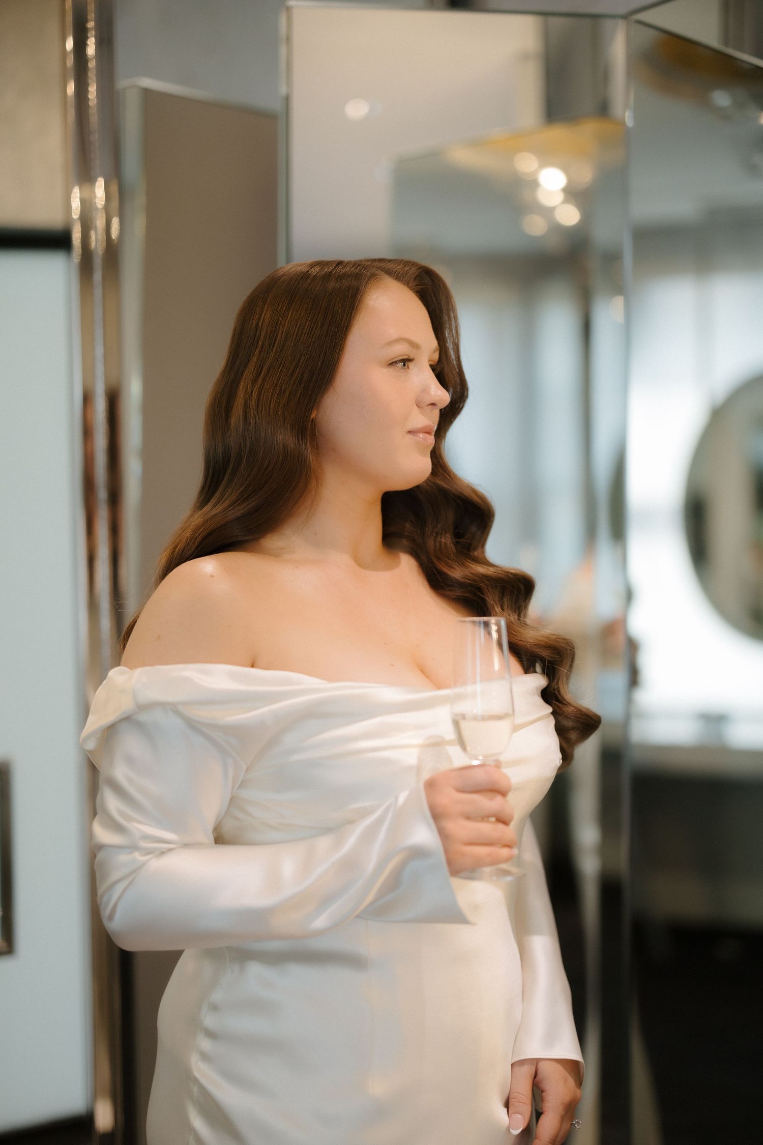 Bride in white off-shoulder satin dress holds champagne glass while gazing sideways in a luxurious dressing room.