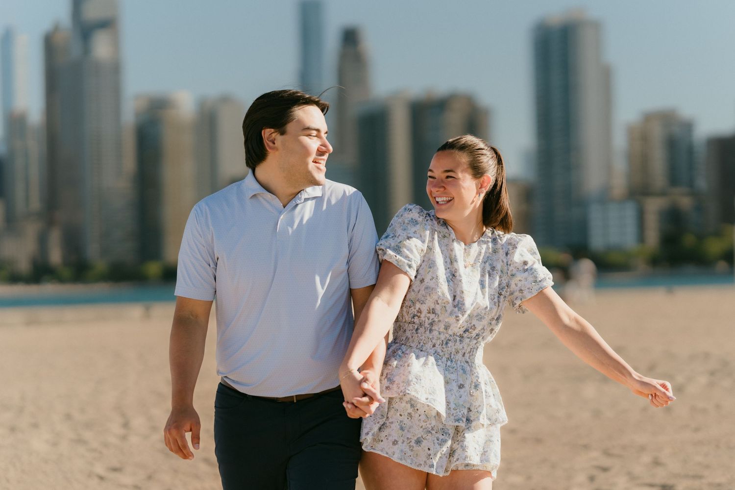 Joyful couple holding hands and laughing on Chicago beach with city skyline behind them.
