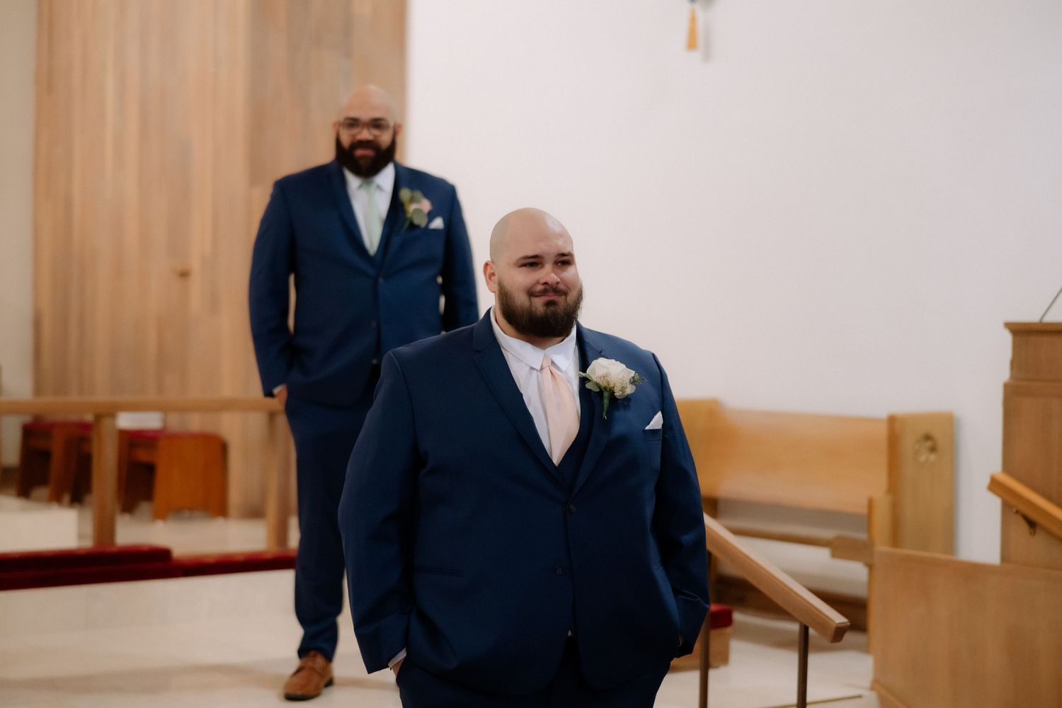 Two groomsmen in navy blue suits with boutonnieres stand in a church aisle before a wedding ceremony.