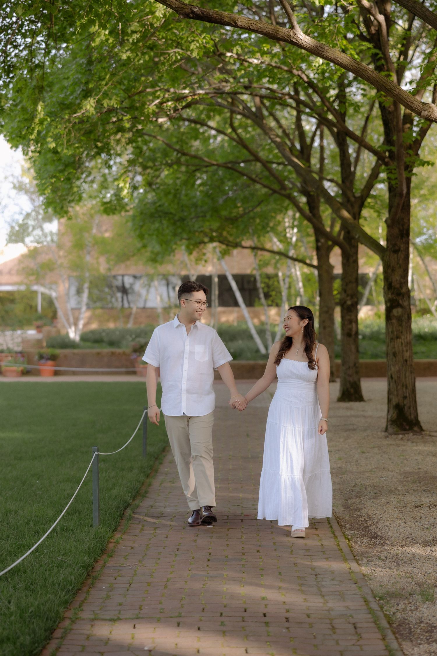 A couple in white attire holds hands while walking along a brick path in a lush green park.