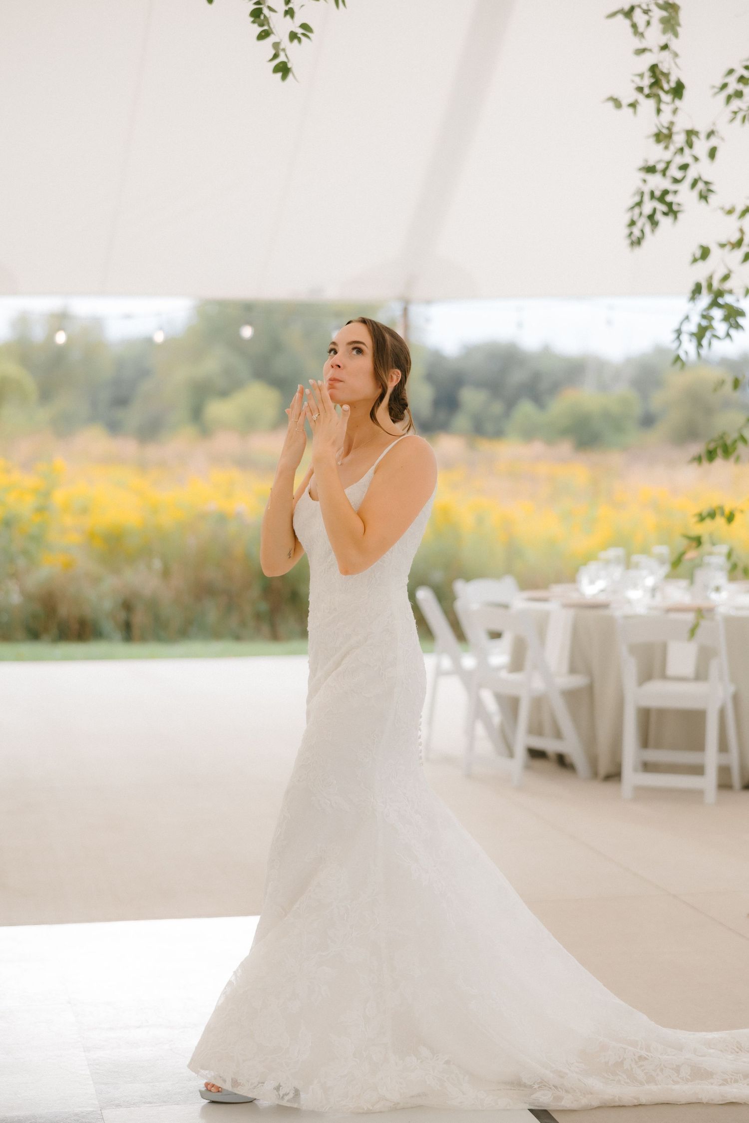 Bride in elegant lace mermaid gown standing in tent reception venue with yellow wildflower field view.