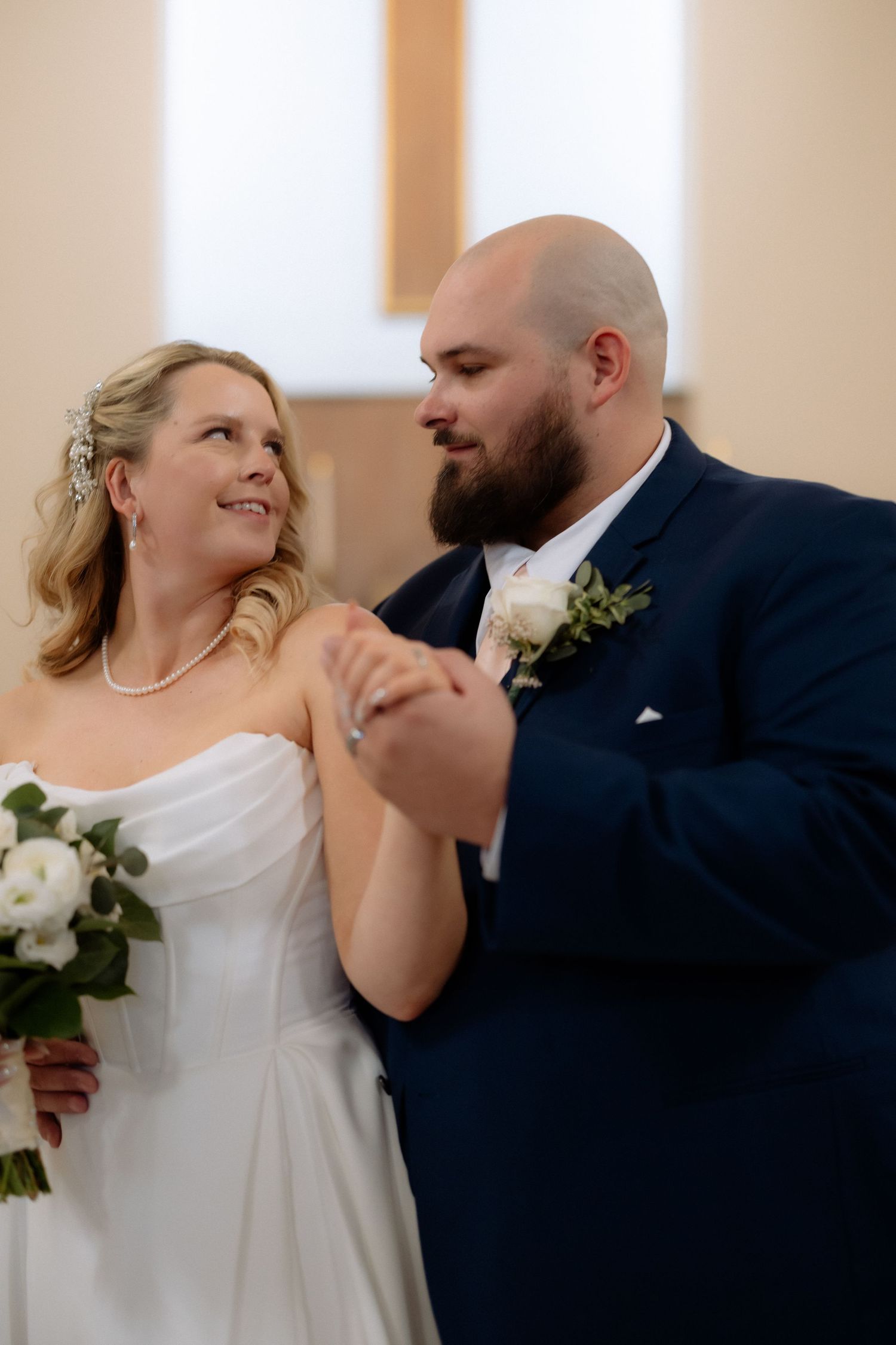 Bride and groom share first dance in church, she holds white bouquet, he wears navy suit with boutonniere.