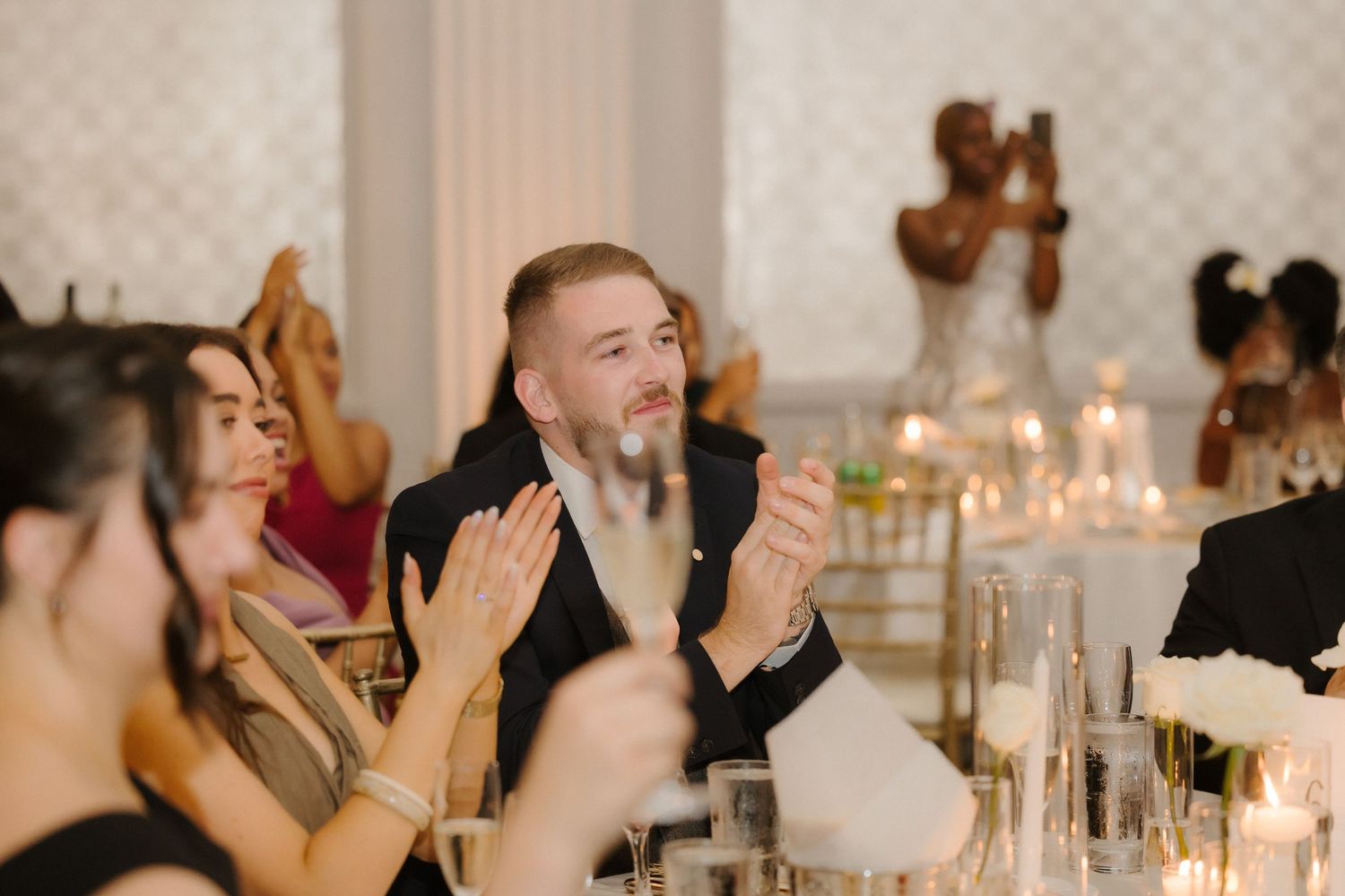 Guests clapping and smiling at an elegant wedding reception with candles and white decor.