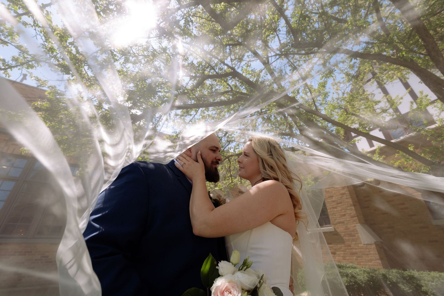 Bride and groom share a romantic moment under a flowing veil with sunlit trees in the background.