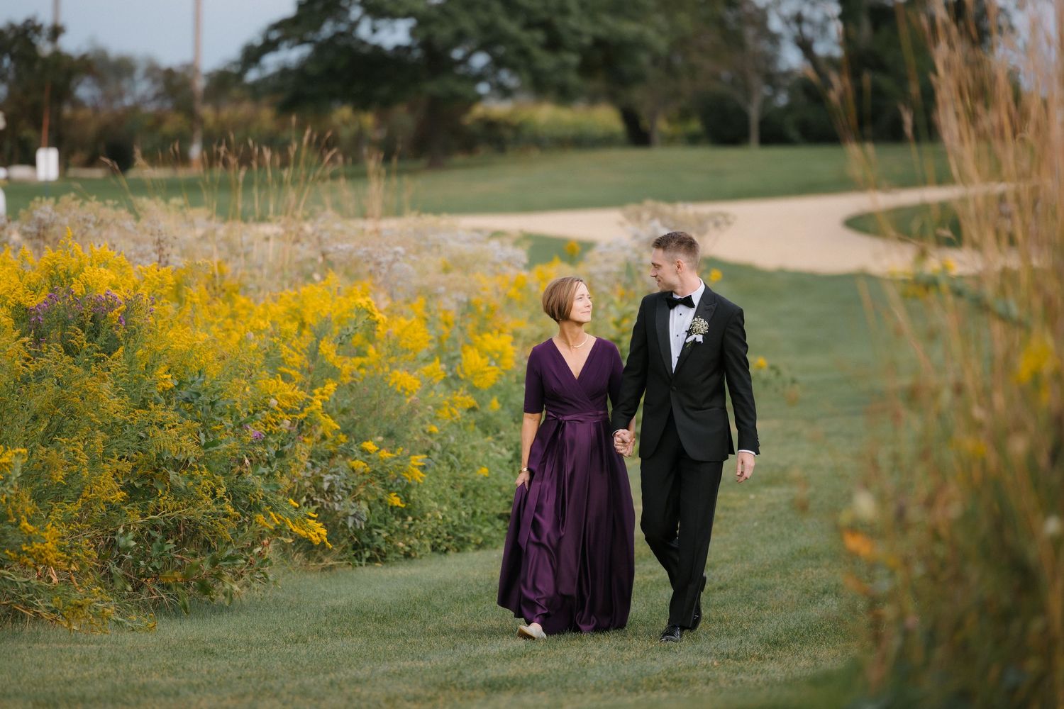 Groom in tuxedo walks with woman in purple gown along path bordered by yellow wildflowers.