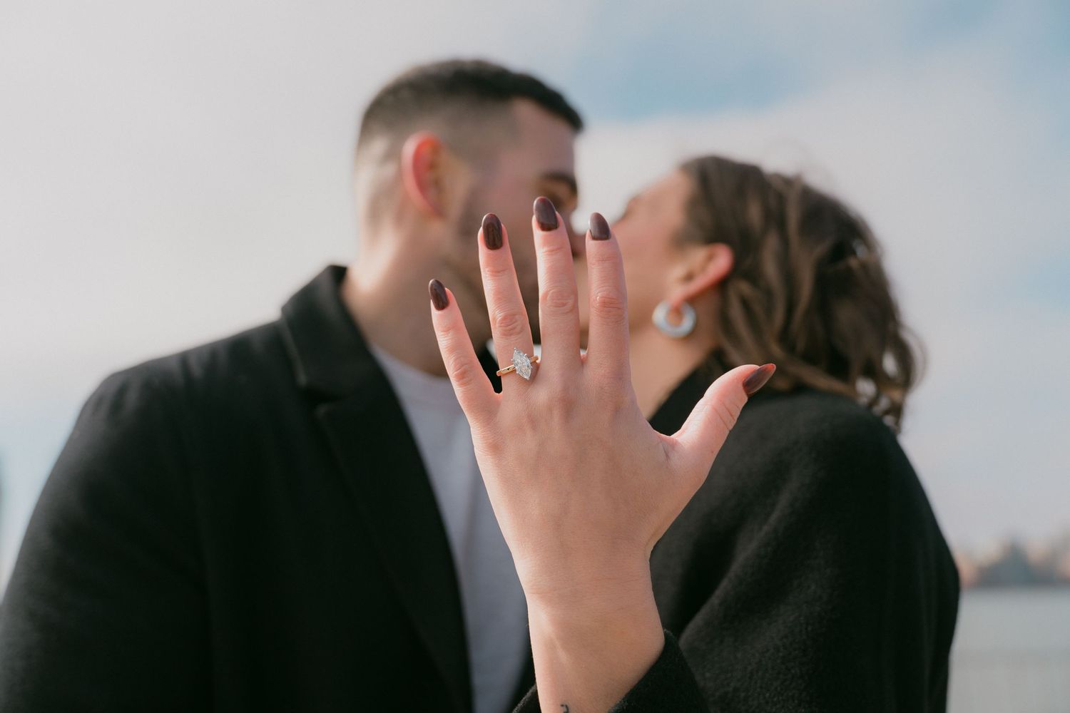 Close-up of sparkling diamond engagement ring on womans hand as couple shares romantic kiss outdoors.