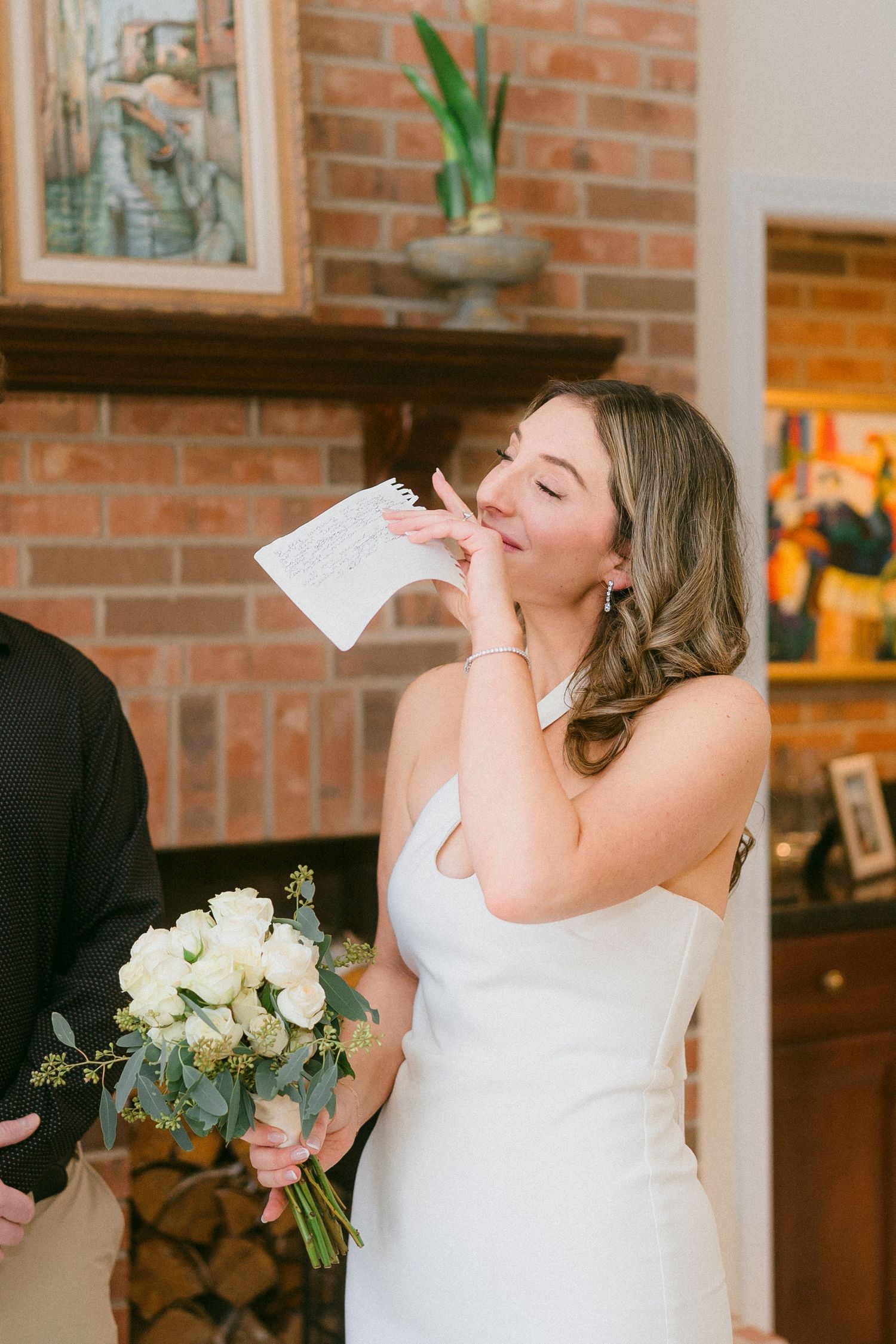 Emotional bride in white gown kissing vow notes while holding white rose bouquet.