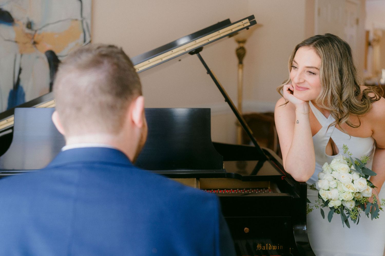 Bride smiling warmly at groom playing grand piano, holding white floral bouquet at wedding venue.