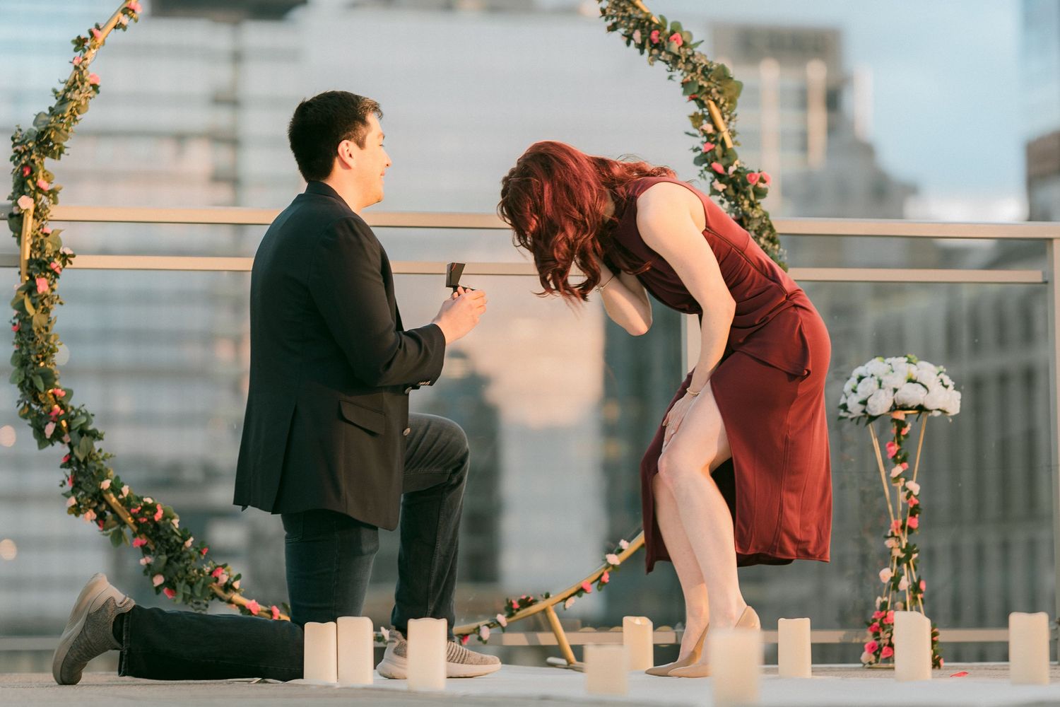 Close-up of man kneeling presenting engagement ring to emotional woman on a rooftop with city views.
