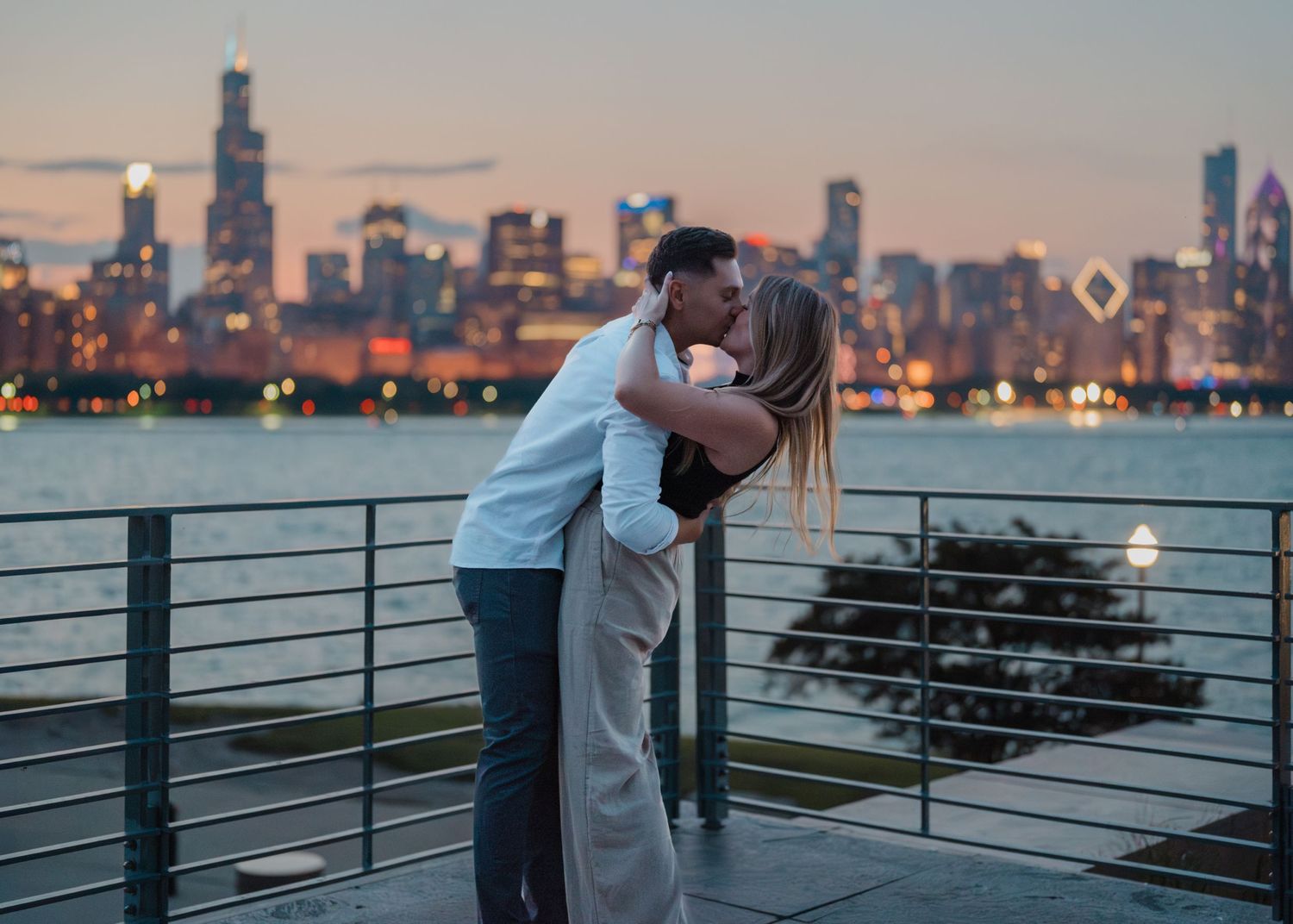A couple shares a passionate kiss on a lakeside deck with the illuminated Chicago skyline at twilight.