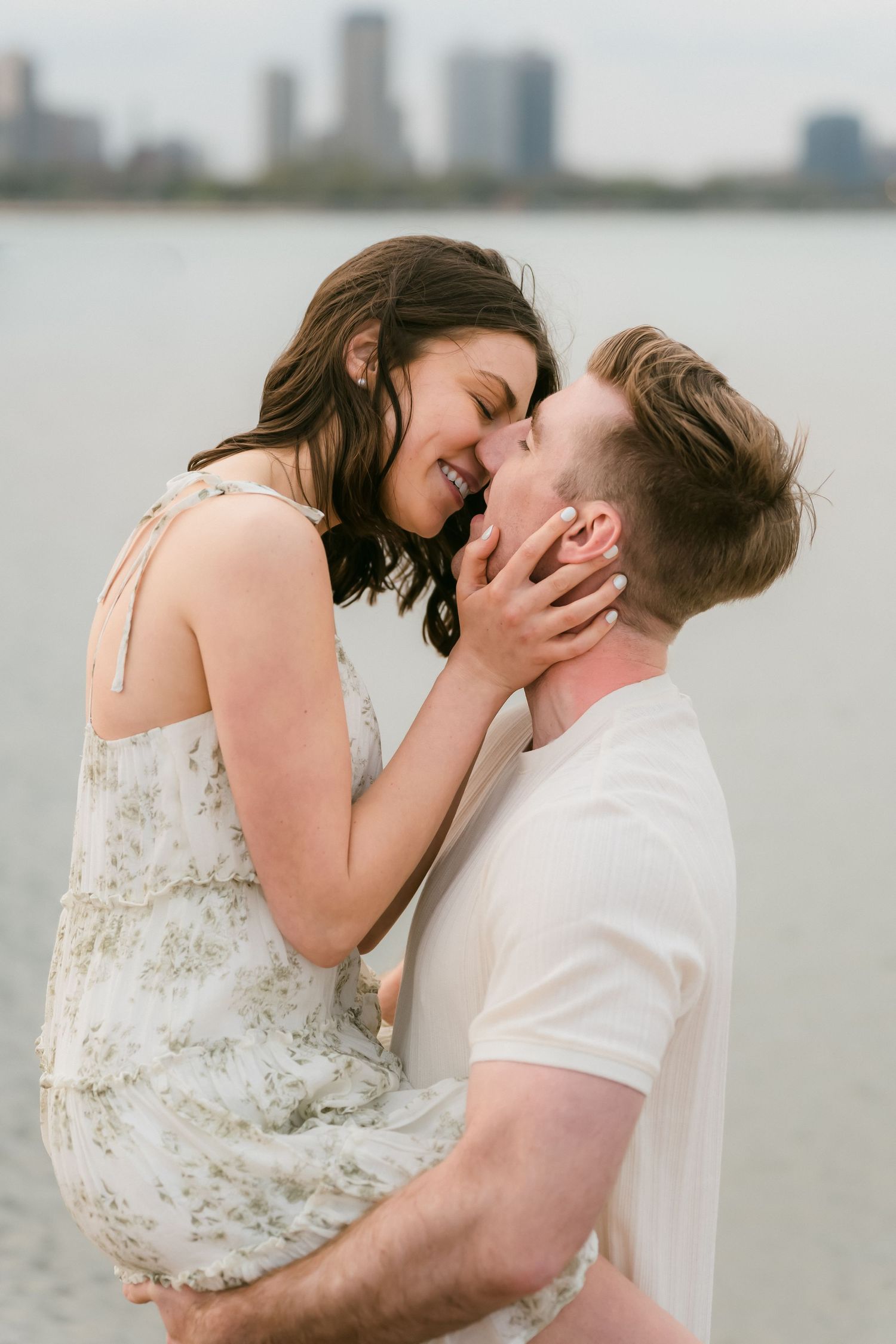 Romantic couple sharing an intimate moment, woman in floral dress being lifted by man near a city waterfront.