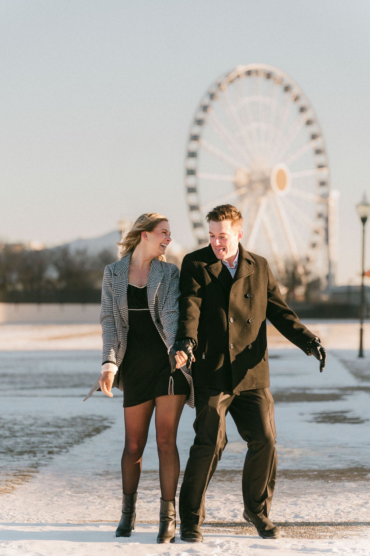 A joyful couple walks hand-in-hand in the snow near a Ferris wheel during golden hour.