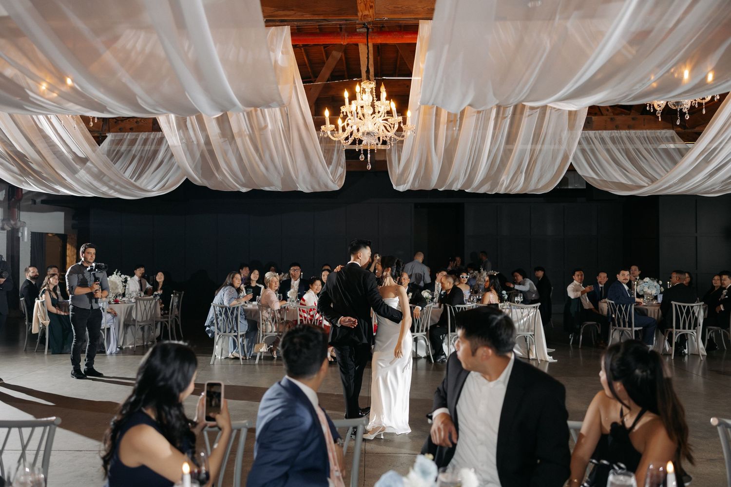 Elegant wedding reception with couple sharing first dance under draped white fabric ceiling and chandelier lighting.