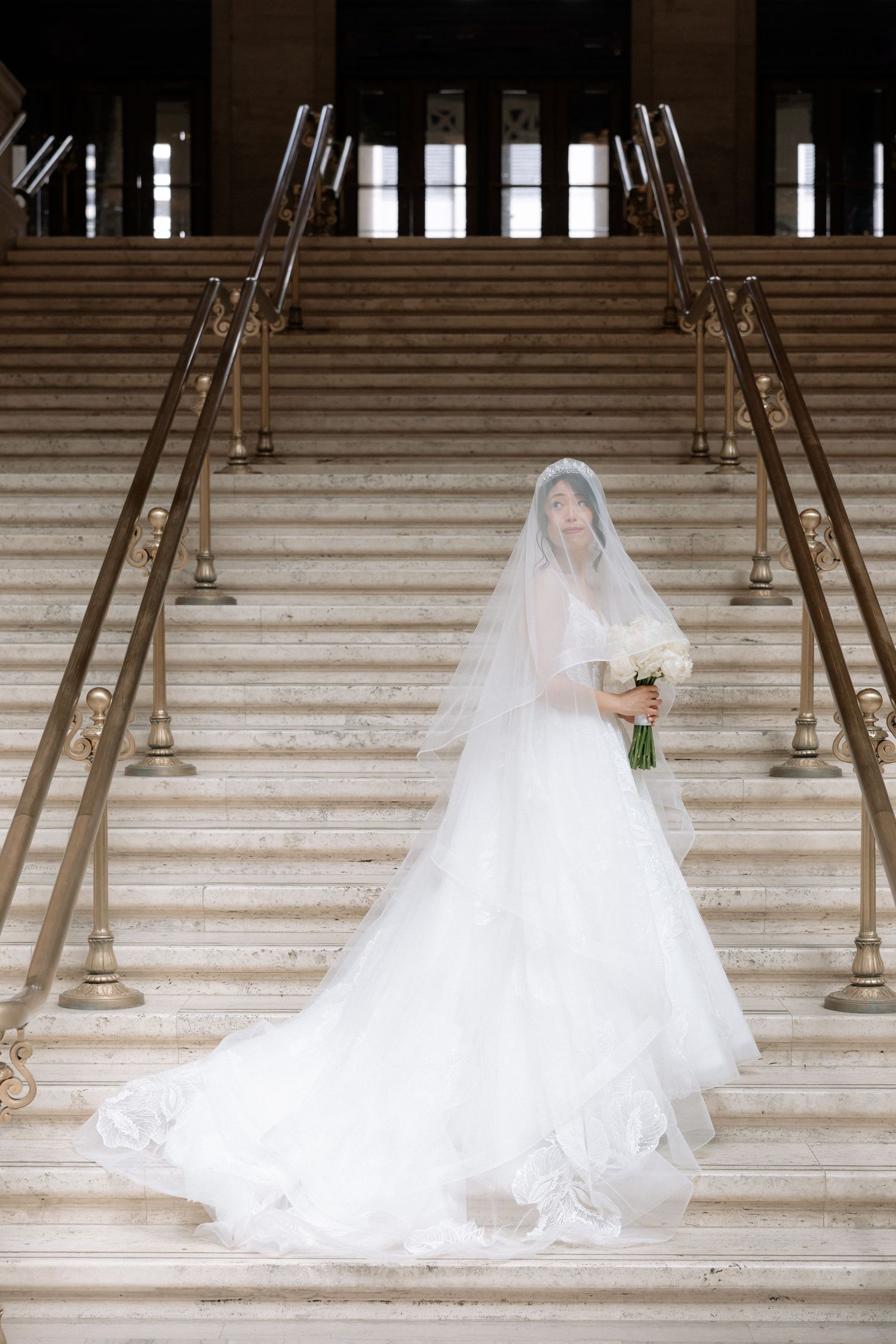 Bride in full white ballgown and long veil holds bouquet while posing on marble staircase in grand venue.