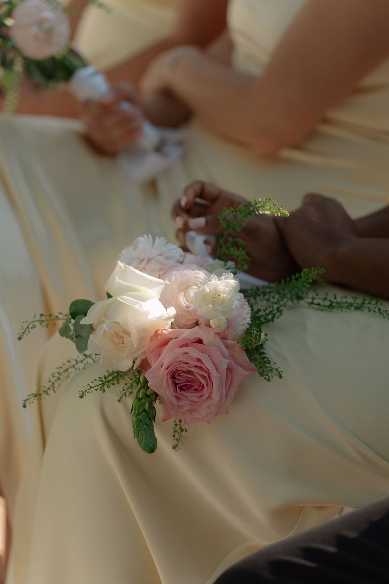 Bridesmaids in cream dresses holding delicate pink and white rose wrist corsages with greenery at a wedding ceremony.