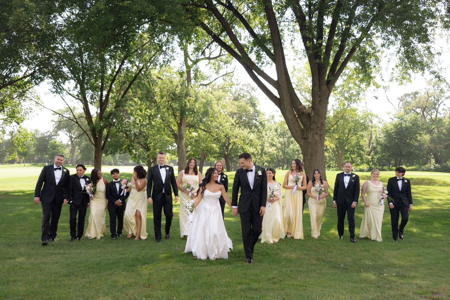 Wedding party walks through park, bride and groom leading with bridesmaids in gold dresses.