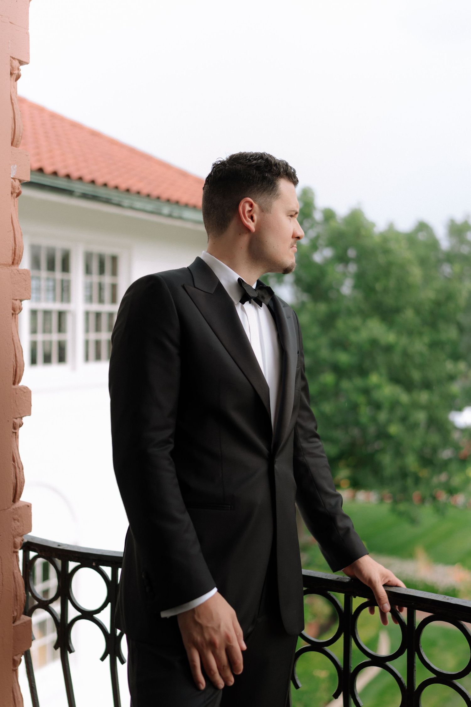 Elegant groom in black tuxedo and bow tie stands on ornate balcony of white wedding venue.