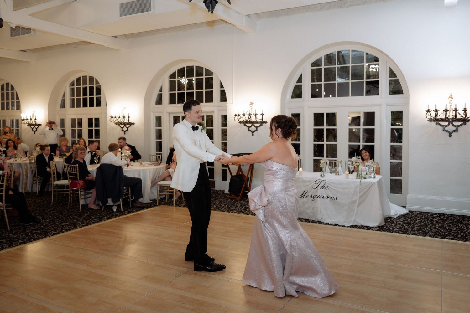 Bride and groom share first dance in elegant ballroom with arched windows and chandeliers.