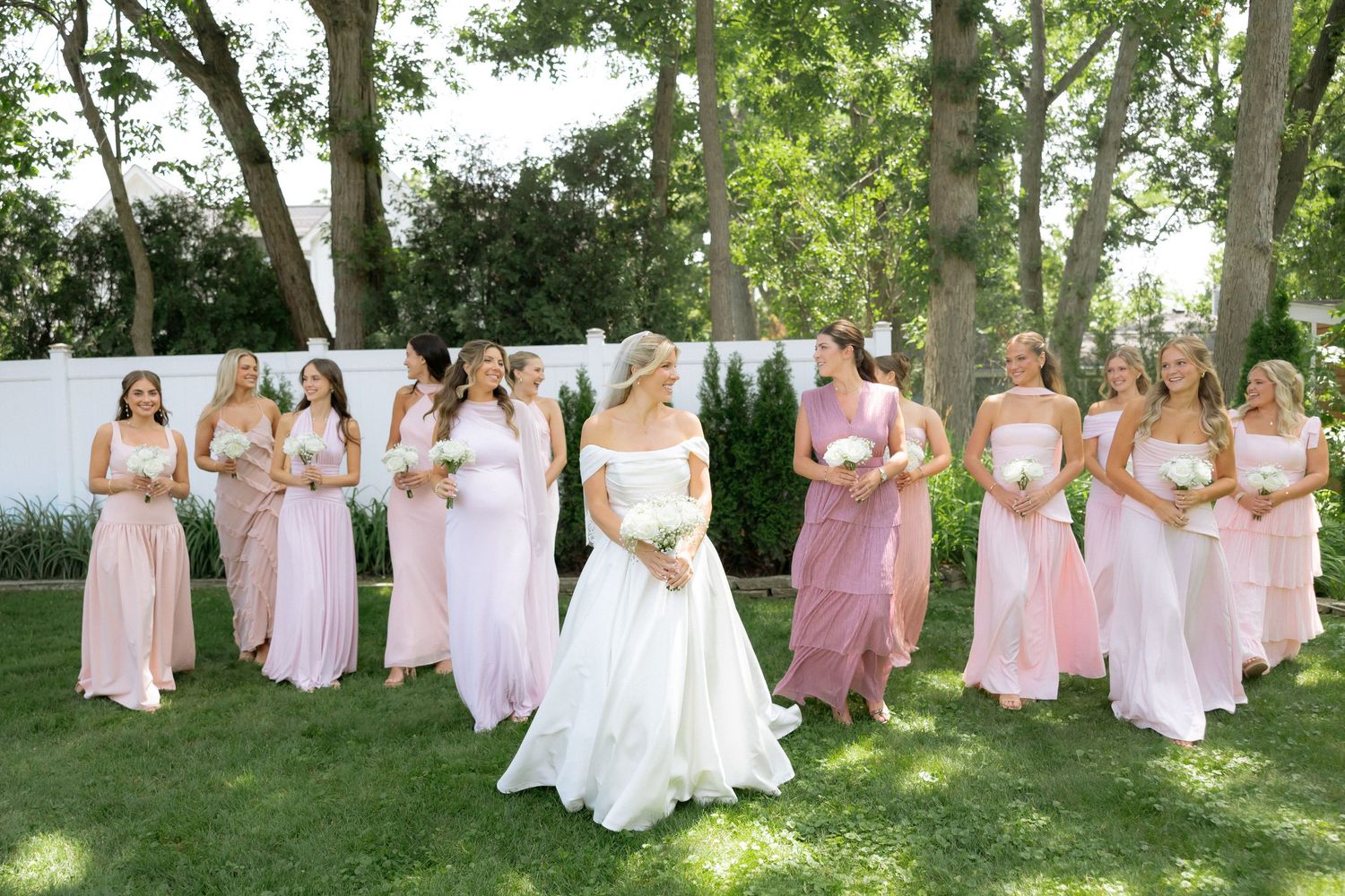 Bride in white ball gown poses with bridesmaids in pink dresses on lush green lawn.