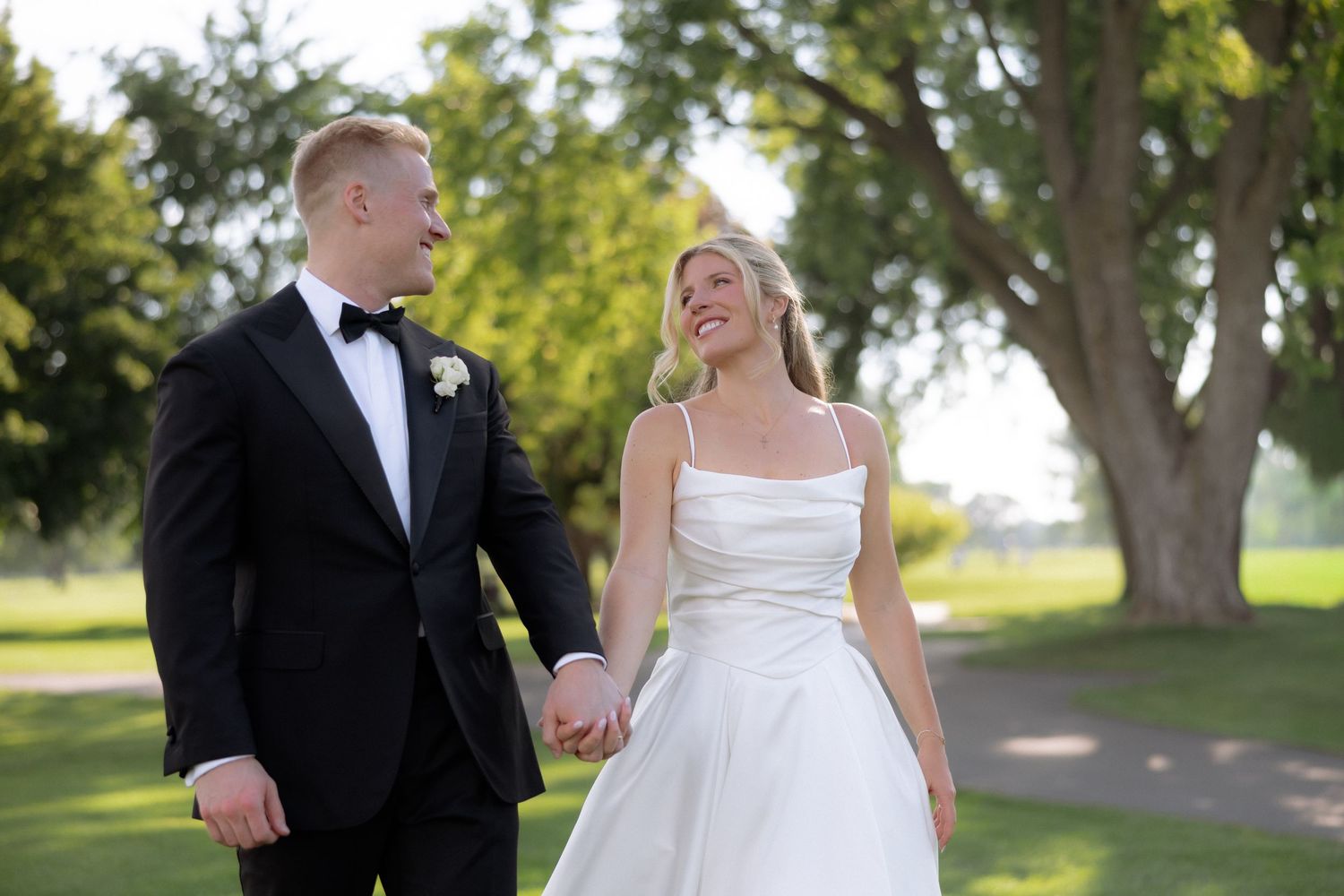 Bride and groom holding hands walking outdoors on golf course greenery on their wedding day.
