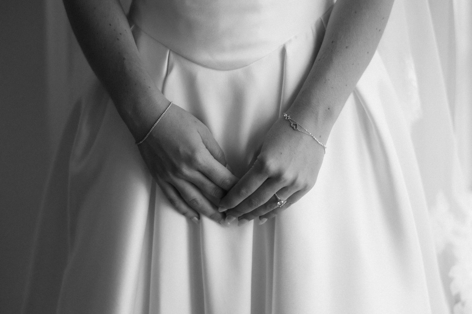 Black and white photo of a bride in a white wedding dress with clasped hands wearing a delicate bracelet.