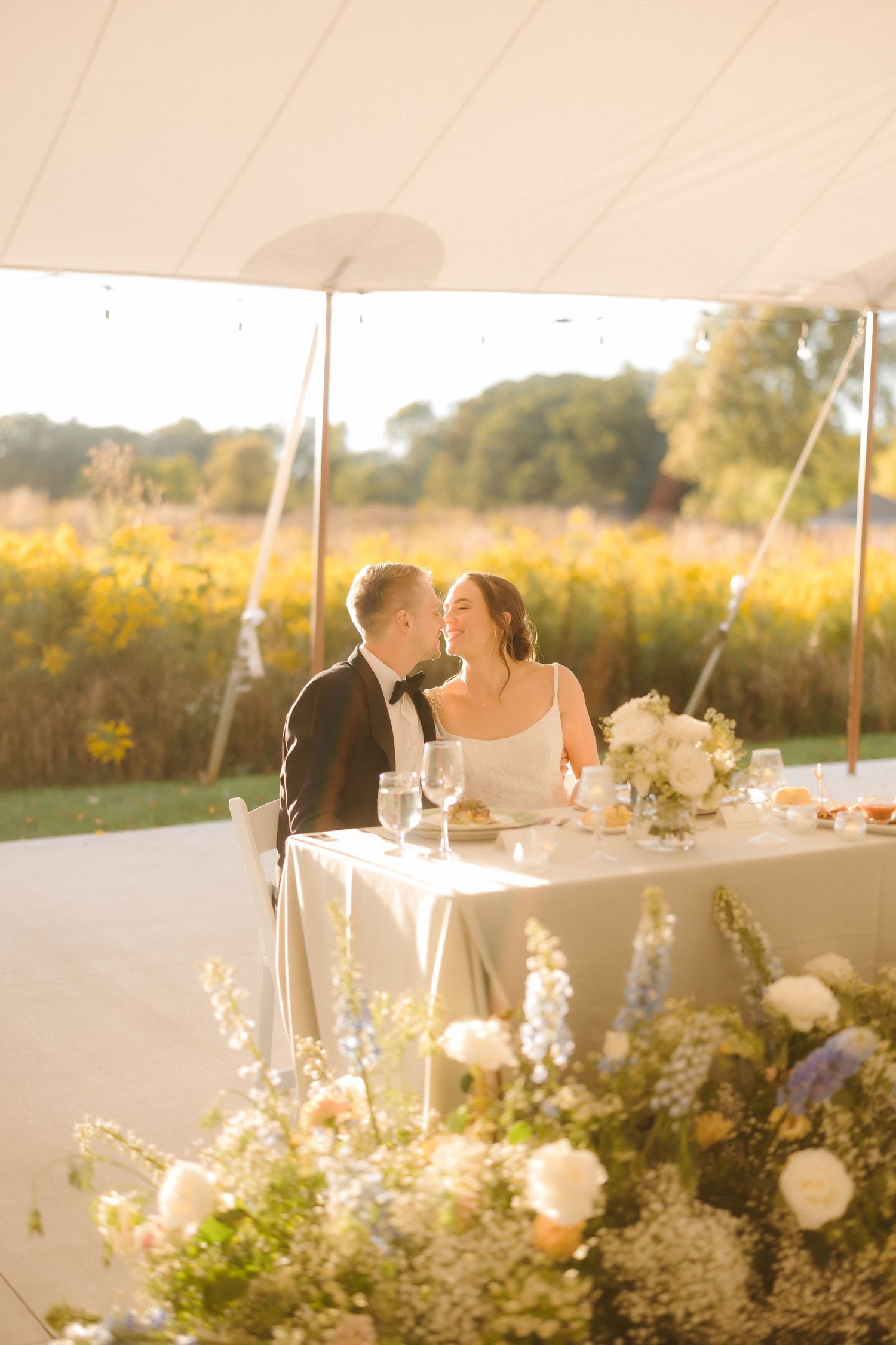 Bride and groom share a romantic moment at sweetheart table under tent, surrounded by wildflowers in golden hour light.