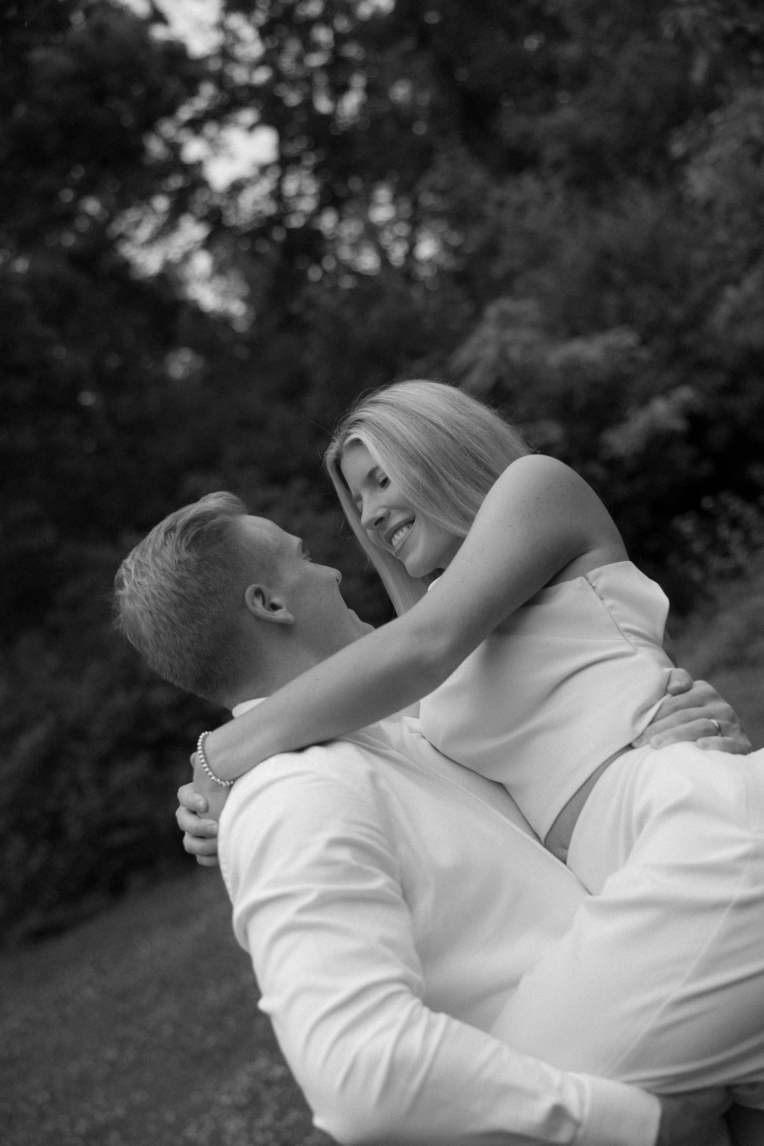Black and white romantic photo of a man lifting a smiling blonde woman in an outdoor park setting.
