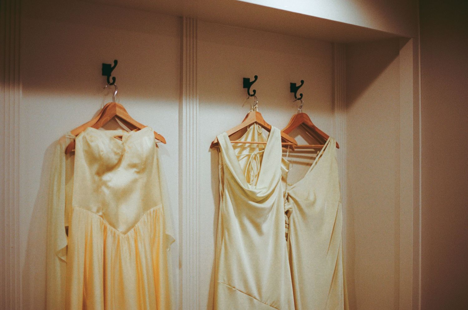 Three elegant cream bridesmaid dresses hanging on wooden hangers with black hooks on a white wall.