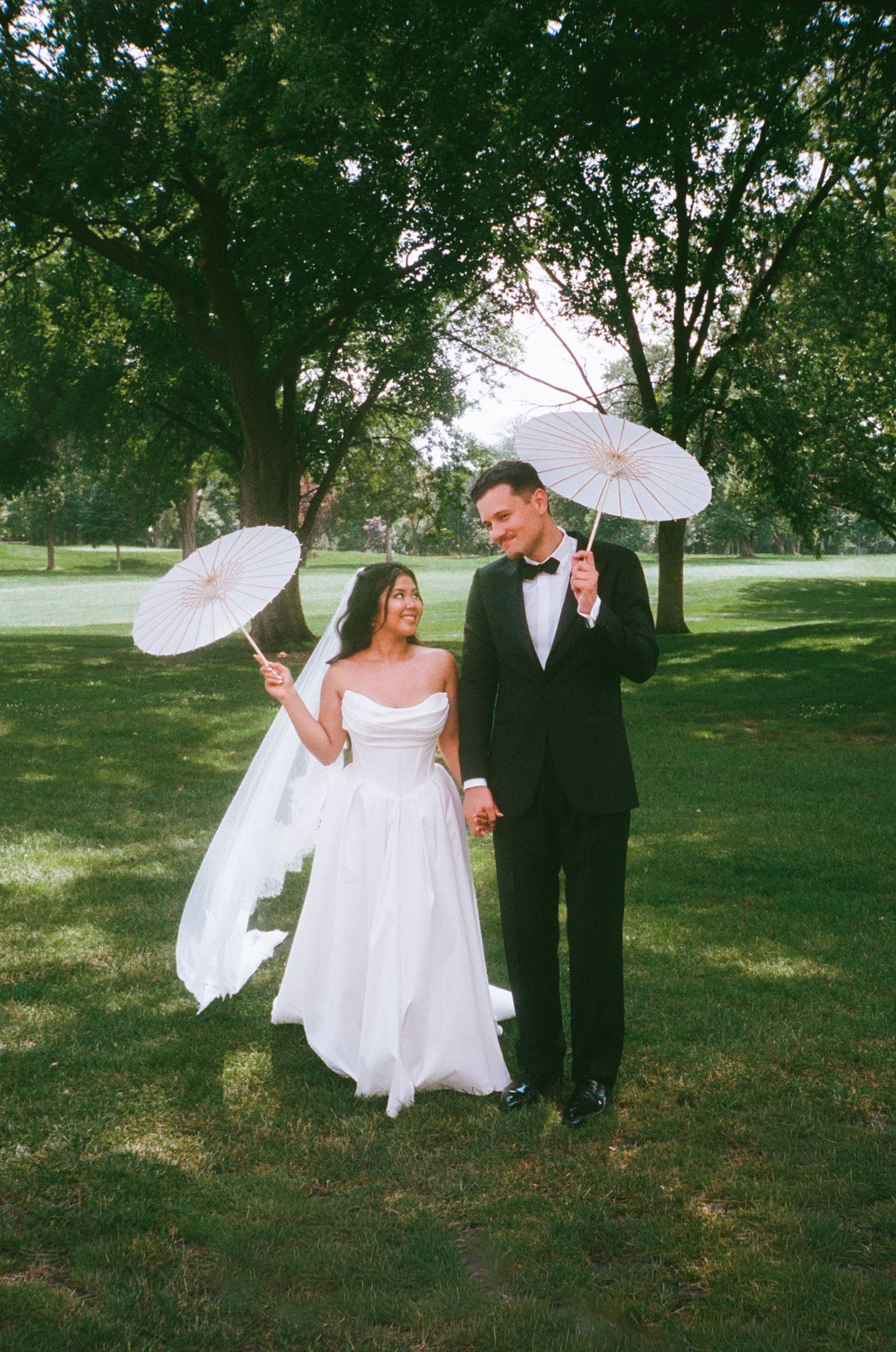 A bride and groom hold white parasols while walking hand-in-hand on a lush green lawn during their outdoor wedding.
