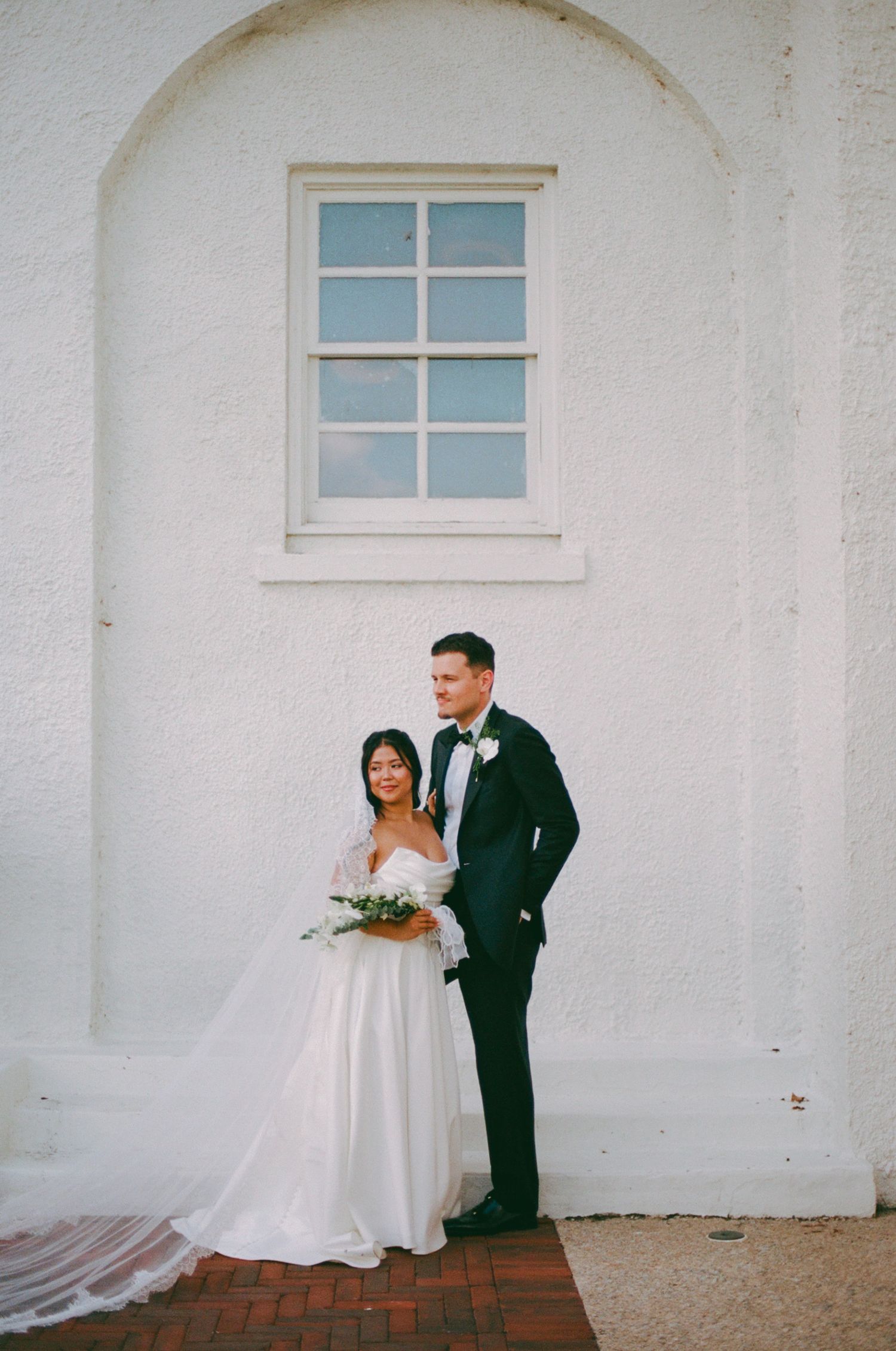 Bride and groom posing elegantly against a white arched wall, bride holding a bouquet in a flowing white gown.