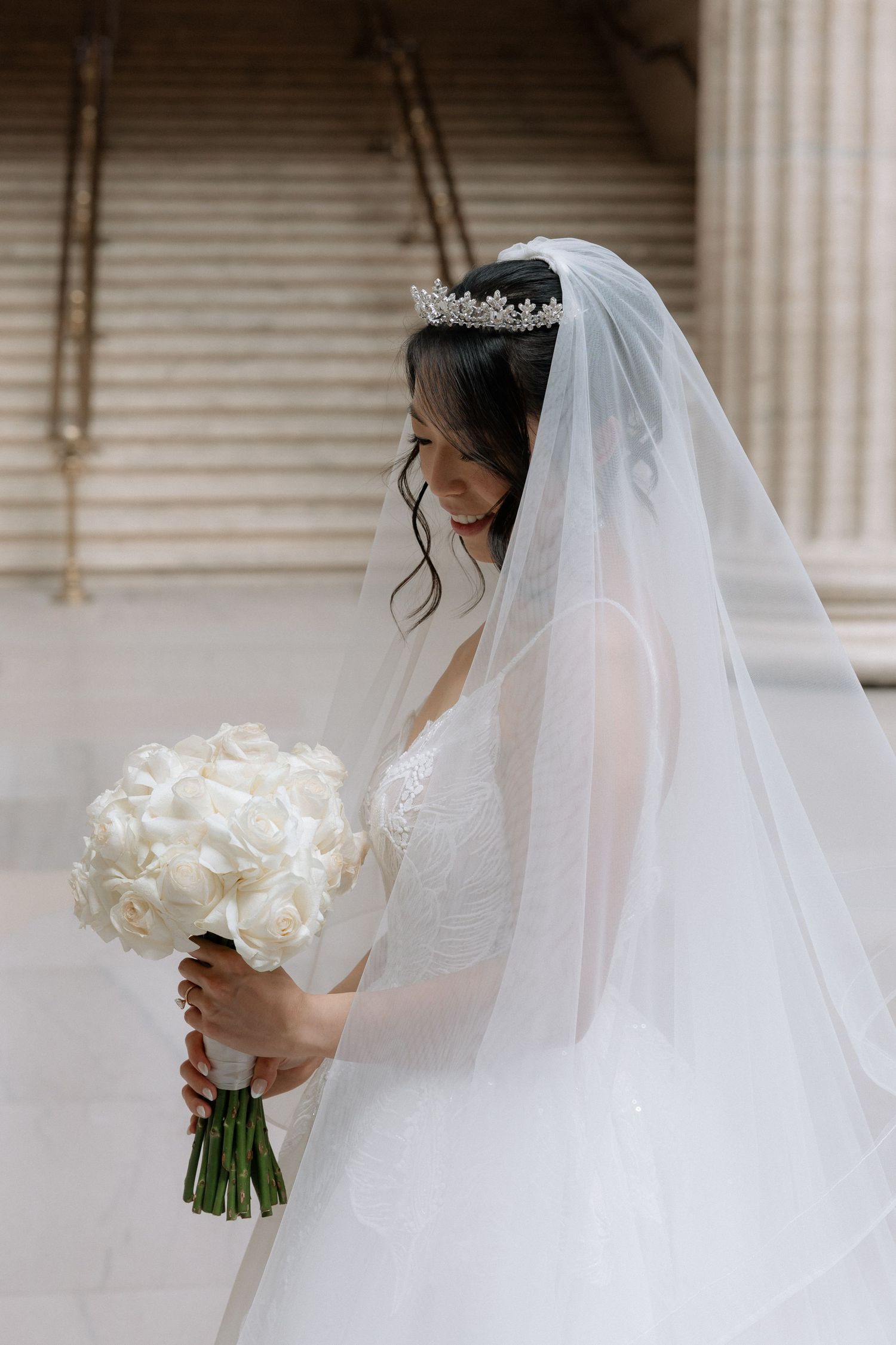 Elegant bride in white lace gown and veil wearing a crystal tiara holds a bouquet of white roses.