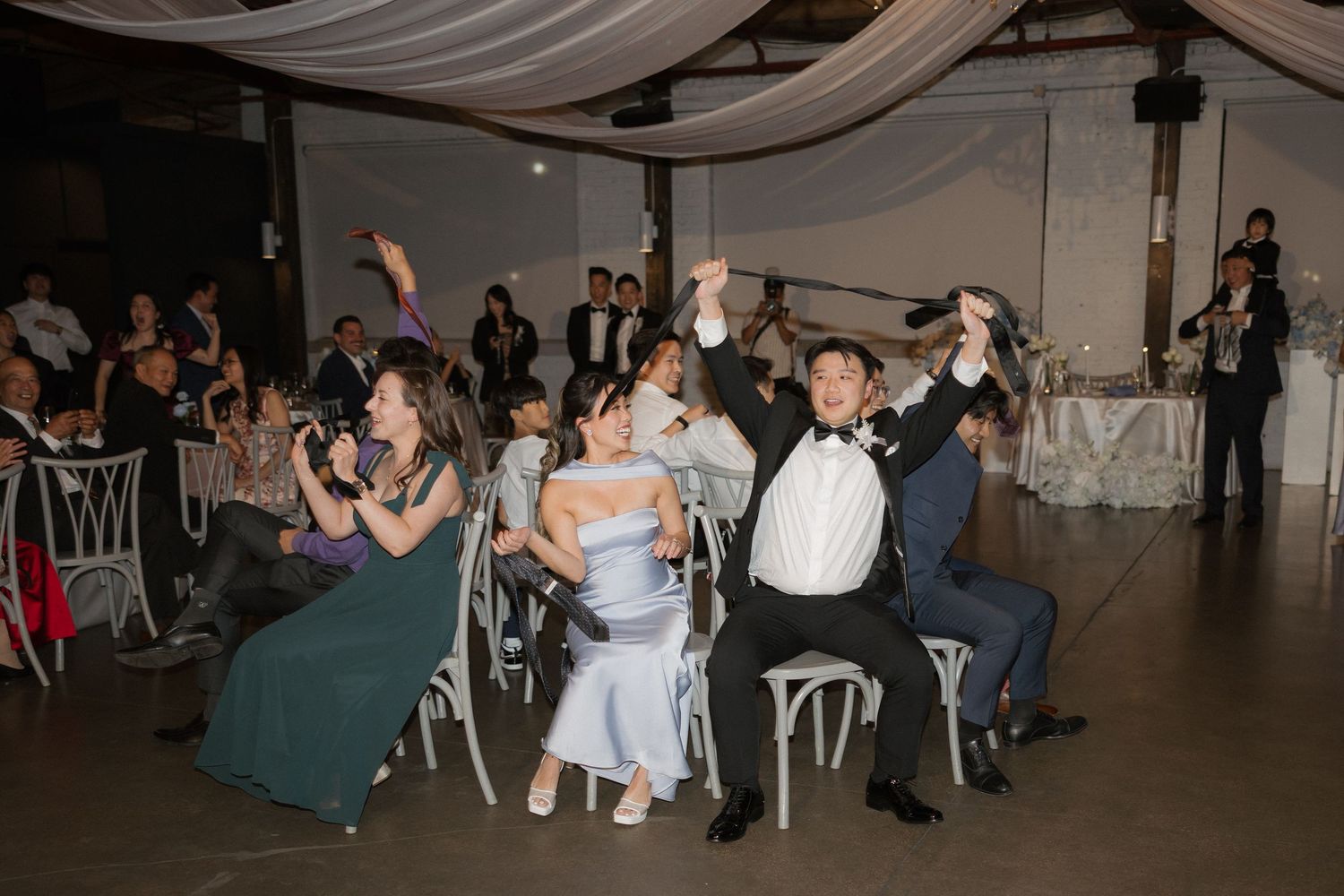Guests celebrate joyfully at a wedding reception, raising hands and clapping in a decorated banquet hall.