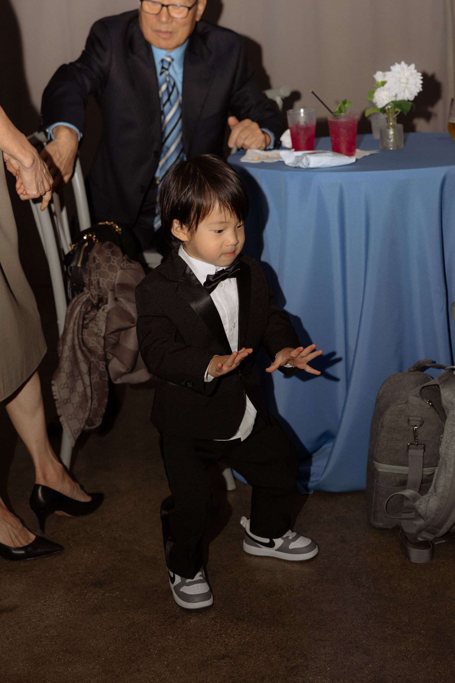 Adorable toddler boy in black tuxedo and bow tie dancing at wedding reception.