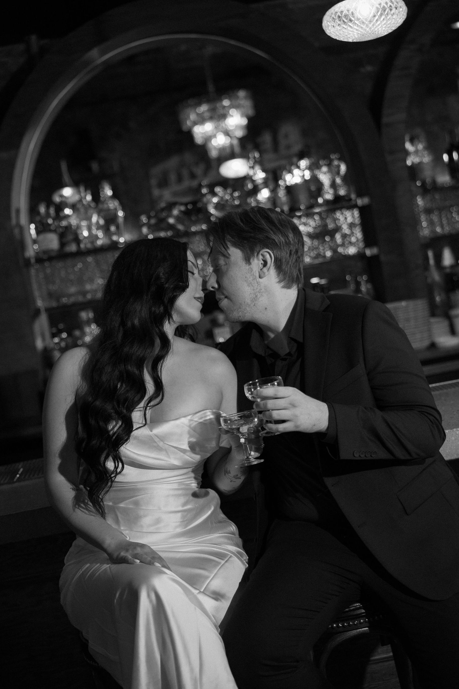 Bride and groom share a kiss at a bar, holding cocktails, in a black and white wedding photo.