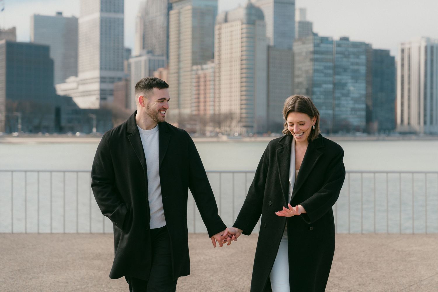 Happy engaged couple holding hands and smiling while walking along Chicago lakefront with city skyline.