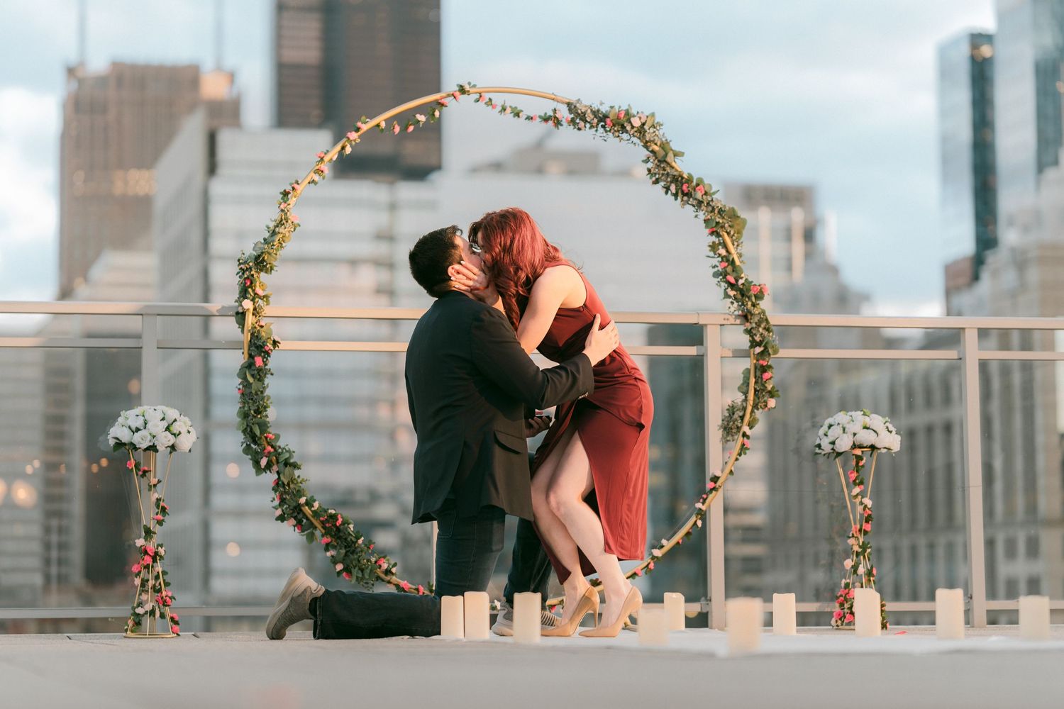 Couple shares first kiss after rooftop proposal framed by a circular floral arch with Chicago skyline behind.