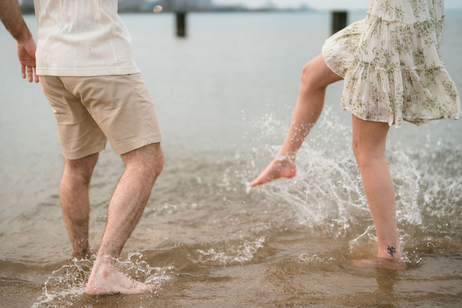 Couple splashing barefoot in shallow water at the beach, wearing casual summer outfits on a cloudy day.