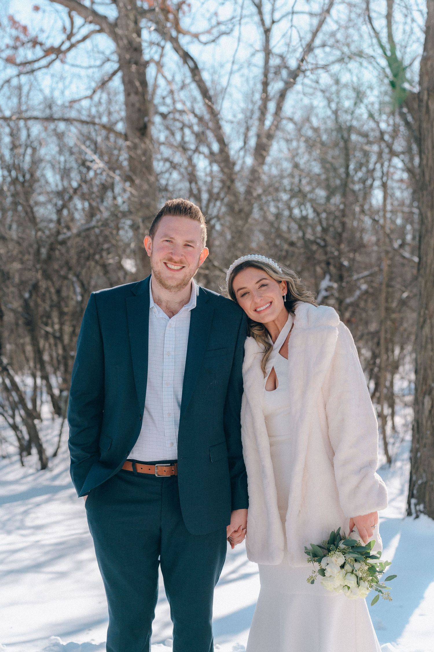 Smiling newlywed couple posing together in snowy winter setting, bride holding white bouquet.